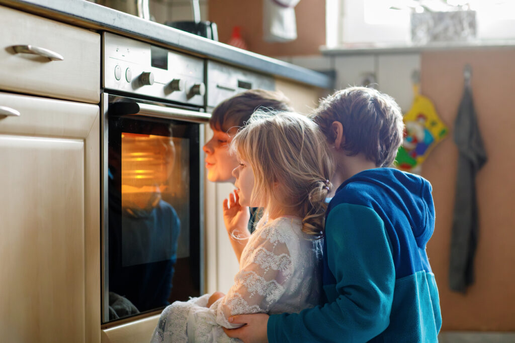 Little toddler girl and two kids boys baking pumpkin cake or cookies in kitchen. Three children, siblings, brothers and sister sitting near oven and waiting. On thanksgiving or christmas holiday.